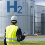 Ingeniero industrial con casco y chaleco reflectante observa una planta de almacenamiento de hidrógeno, con tanques metálicos marcados con “H₂”.