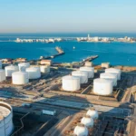 Aerial view of an aboveground oil storage terminal with multiple tanks and secondary containment systems near a coastal port, illustrating risk-based environmental management and spill prevention.