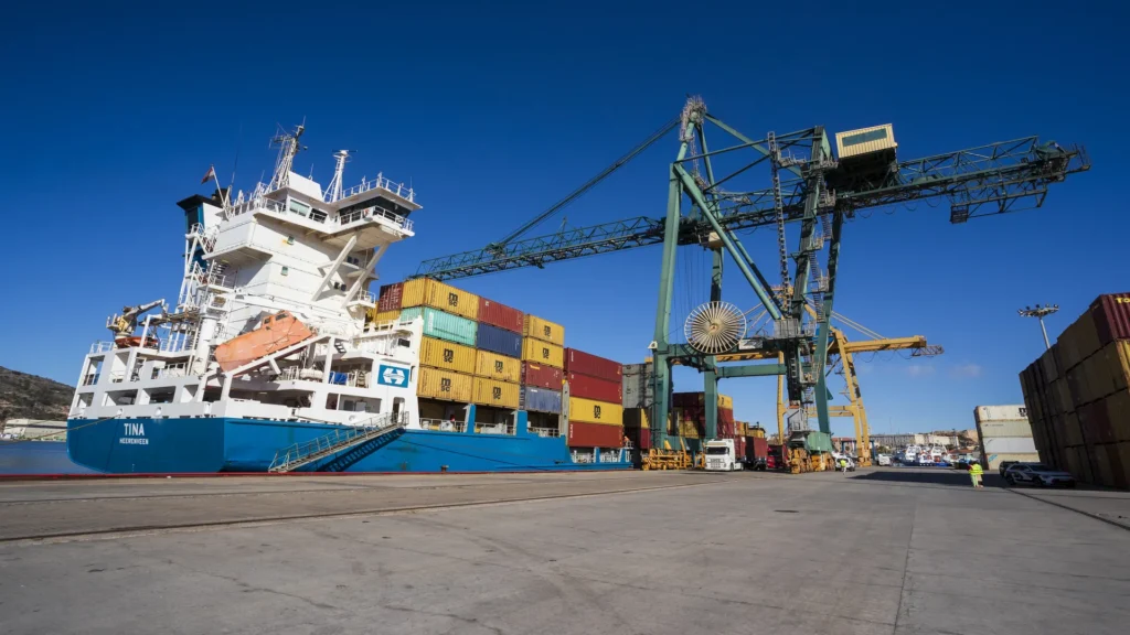 Container ship docked at the Port of Cartagena during loading and unloading operations, with port cranes and containers stacked on the quay under a clear sky.