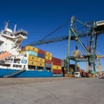 Container ship docked at the Port of Cartagena during loading and unloading operations, with port cranes and containers stacked on the quay under a clear sky.