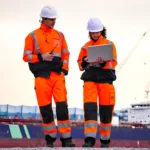 Dos trabajadores con casco y ropa de seguridad naranja revisan información en una computadora portátil mientras caminan en un área operativa de terminales petroleros, con un buque y equipos portuarios al fondo.