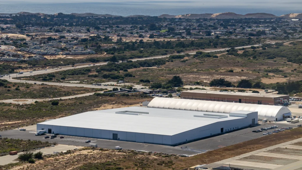 Vista aérea de instalaciones industriales modernas dedicadas a la producción de taxis aéreos eléctricos, con techos blancos, carreteras circundantes y el océano al fondo.