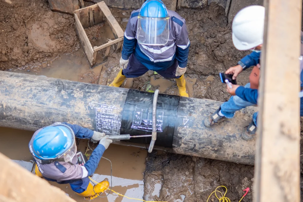 Técnicos con equipo de seguridad inspeccionan una tubería enterrada con fluid-7 en terreno fangoso durante una intervención operativa, marcando datos sobre un recubrimiento compuesto instalado.