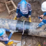 Técnicos con equipo de seguridad inspeccionan una tubería enterrada con fluid-7 en terreno fangoso durante una intervención operativa, marcando datos sobre un recubrimiento compuesto instalado.