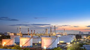 Vista panorámica al atardecer de una instalación industrial con tanques de almacenamiento aéreo (AST), torres de proceso y luminarias encendidas, representando la infraestructura de la industria midstream.