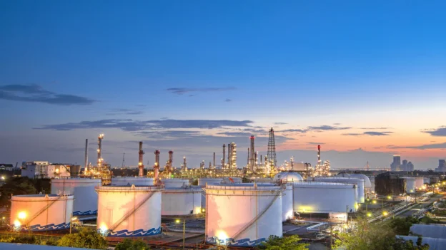 Vista panorámica al atardecer de una instalación industrial con tanques de almacenamiento aéreo (AST), torres de proceso y luminarias encendidas, representando la infraestructura de la industria midstream.