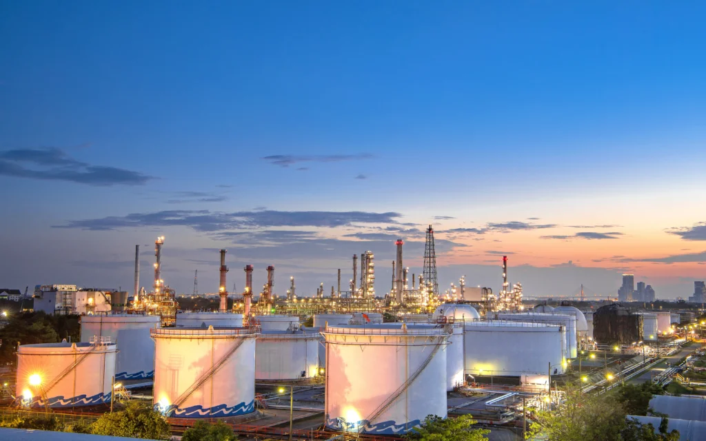 Vista panorámica al atardecer de una instalación industrial con tanques de almacenamiento aéreo (AST), torres de proceso y luminarias encendidas, representando la infraestructura de la industria midstream.