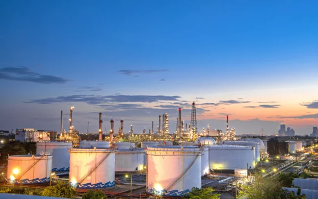 Vista panorámica al atardecer de una instalación industrial con tanques de almacenamiento aéreo (AST), torres de proceso y luminarias encendidas, representando la infraestructura de la industria midstream.