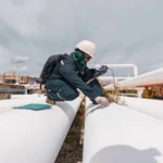Técnico de inspección industrial con EPP realiza medición sobre una tubería blanca expuesta, utilizando un equipo portátil de Ensayo No Destructivo en una planta energética.