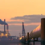 Vista de una planta industrial al atardecer con columnas de humo y una línea de tubería de gran diámetro que se extiende hacia el horizonte, representando infraestructura crítica en la industria de hidrocarburos.
